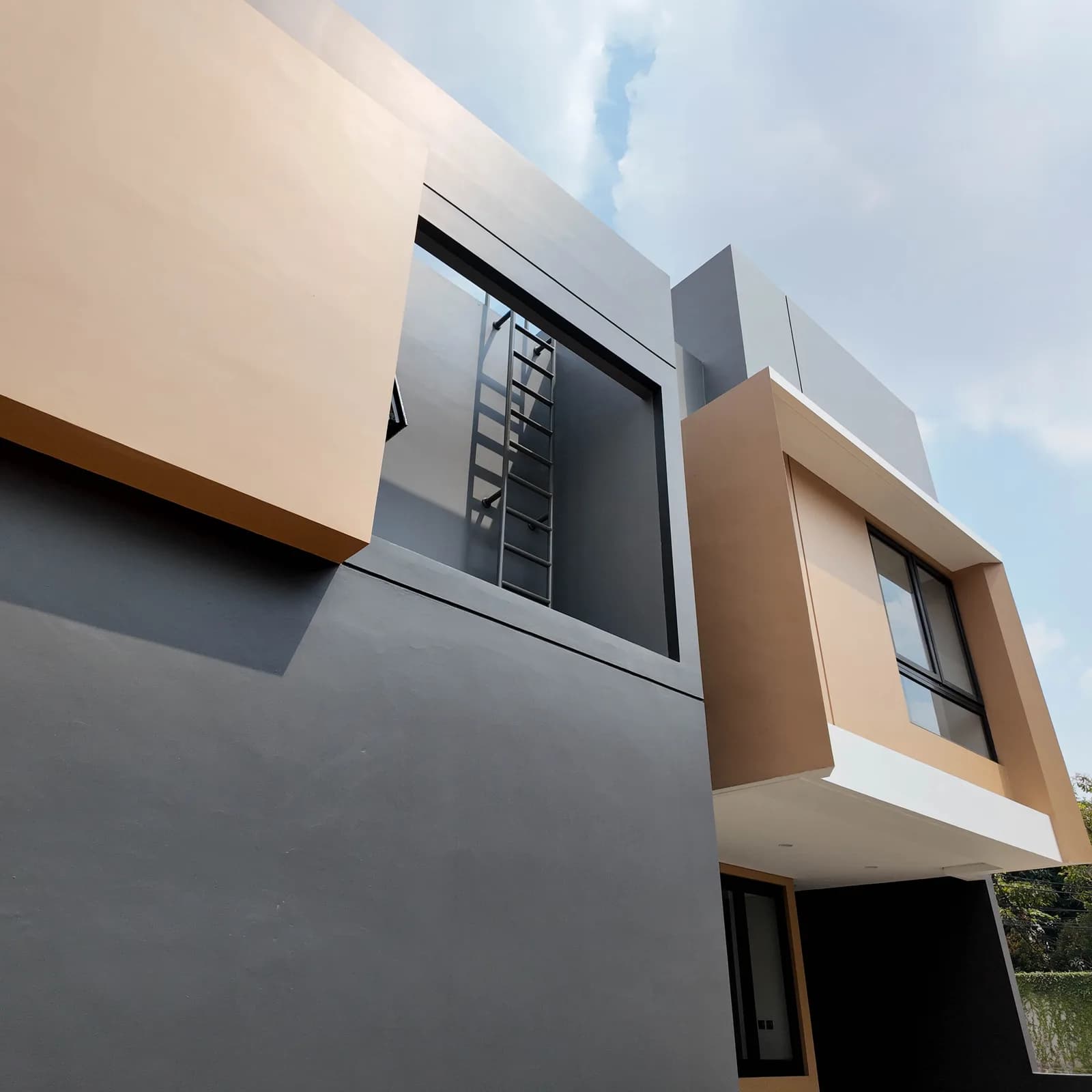 Photograph of a house corner where charcoal and tan-beige wall planes meet, with a roof-access ladder in a recessed shaft