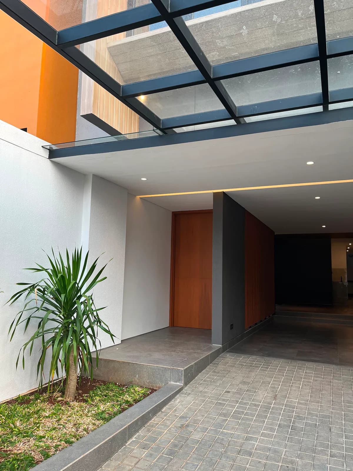 The timber front door set back under a steel and glass canopy, beside the tiled driveway
