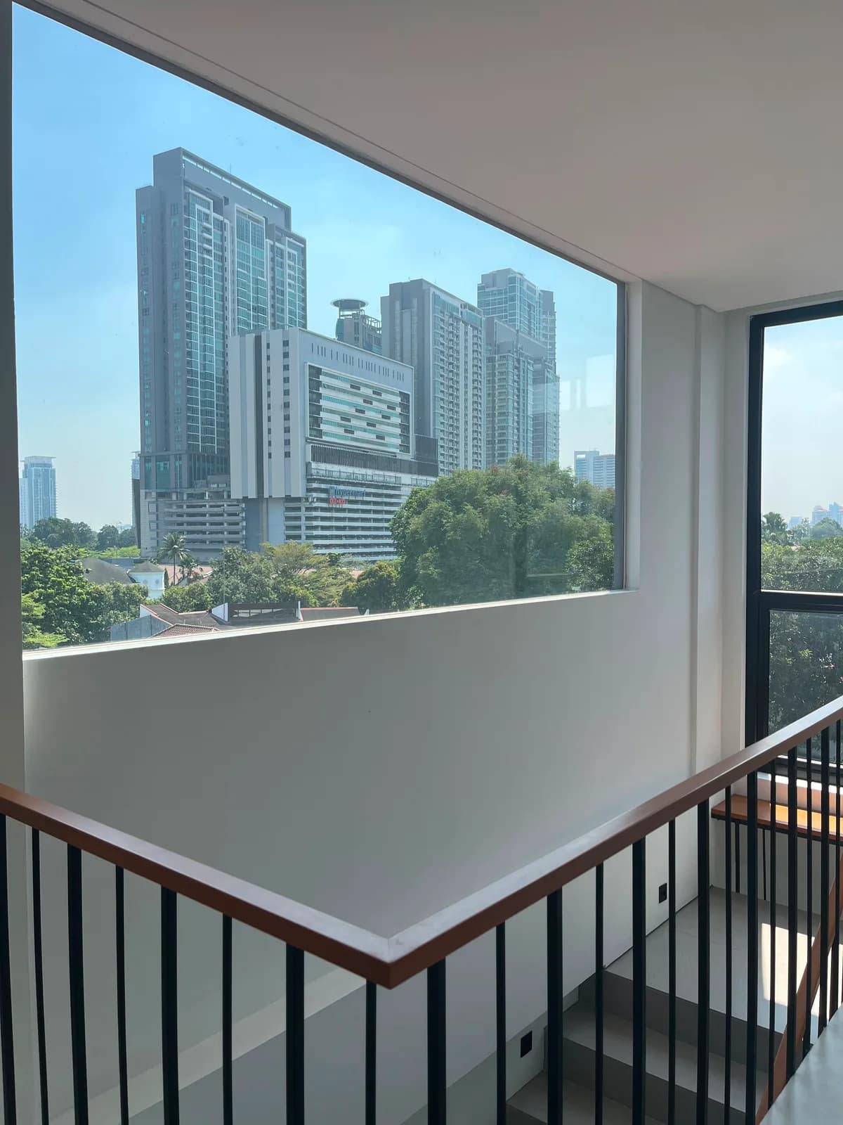A wide window beside the stair void framing the trees and high-rise towers of South Kemang