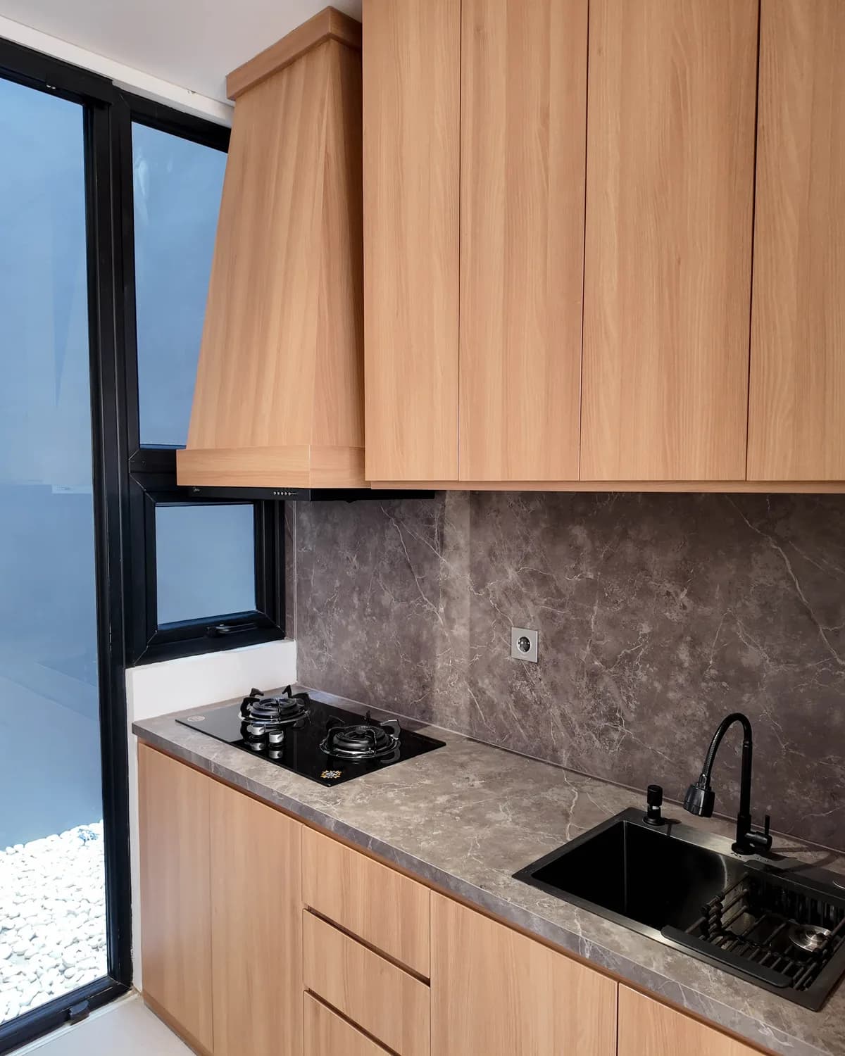 A fitted kitchen with light oak cabinets, a grey marble worktop and splashback, a black gas hob and sink, beside a black-framed glass wall onto a pebbled light court